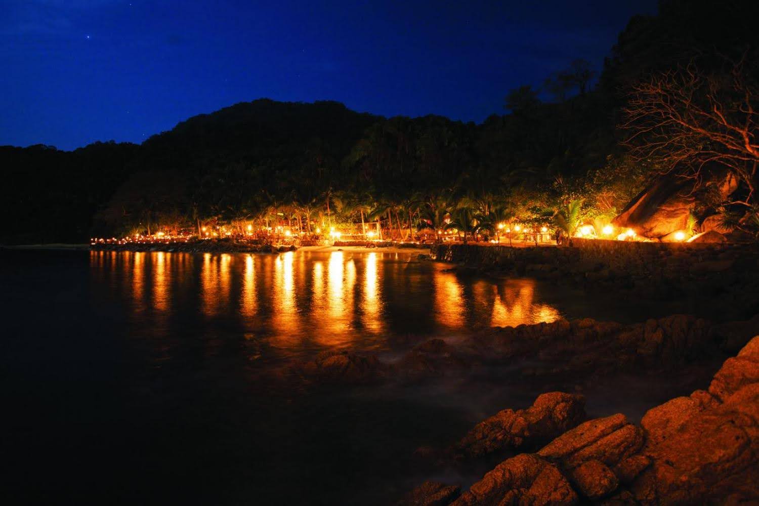 Long exposure night photo of a coastline with warm glowing lights reflected in dark water beneath a deep blue starry sky.